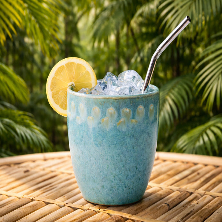 Henkelloser Keramikbecher aus portugiesischer Keramik in blau mit feinen Sprenkeln und hellem Rand gefüllt mit Strohalm und Eiswürfeln in Naturumgebung